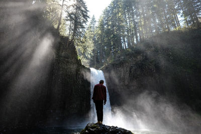 Rear view full length of man standing on rock against waterfall at forest