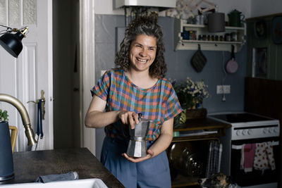 Happy woman holding coffee pot while standing in kitchen at home