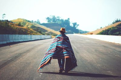 Rear view of man with blanket walking on road against sky