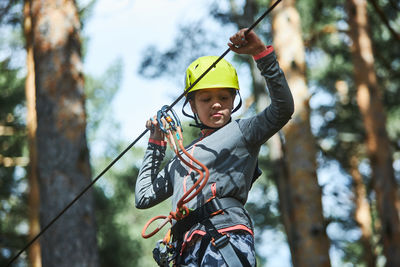 From below girl in activewear and helmet with safety ropes grasping line and balancing on obstacle course on blurred background of coniferous trees in woods