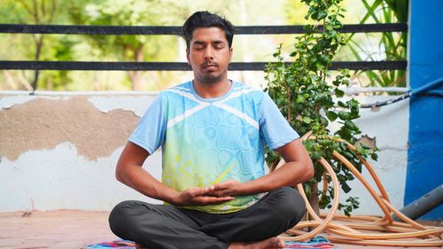 Full of joy young indian man wearing sports clothes blue shirt and black tights in lotus position
