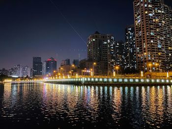 Illuminated buildings by river against sky at night