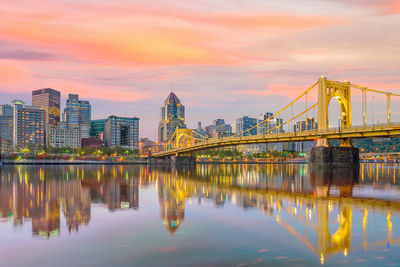 Bridge over river by buildings against sky during sunset