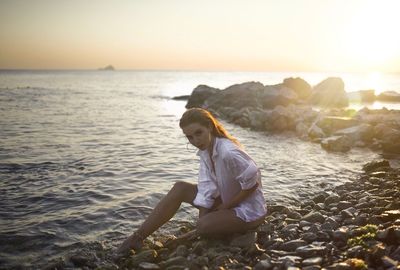 Man sitting on rock at beach against sky during sunset