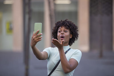 Happy female blogger with afro hair holding phone having video call while walking in the city