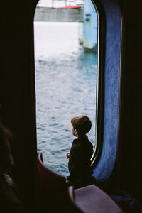 Rear view of boy looking through window of ship on sea