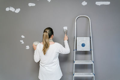 Backwards blonde woman plastering with a spatula a wall after being painted with a grey painting. the painter is next to her ladder while covering small holes on the wall. horizontal picture