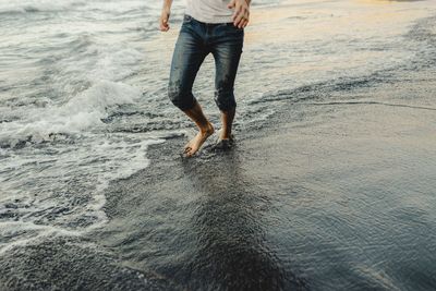 Crop anonymous barefoot male traveler in jeans walking on sandy beach near waving sea at sunset time