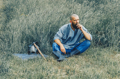 Courageous man with beard resting in grass in field. pensive gaze. next to him is ax