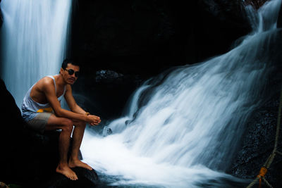 Young man sitting on rock against waterfall
