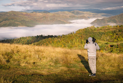 Man taking photos standing on field against foggy mountains and sky
