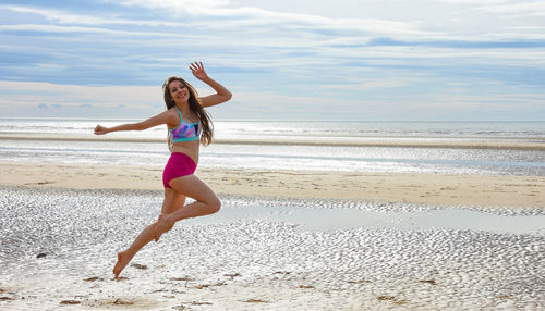 Full length portrait of young woman jumping at beach against sky