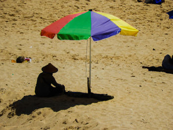 Rear view of man wearing hat sitting under umbrella at beach