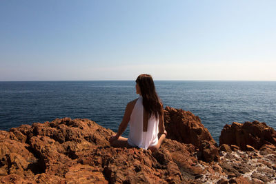 Rear view of woman sitting on rock by sea against clear sky