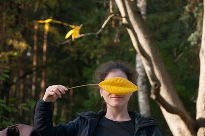 Portrait of woman holding yellow leaf against trees