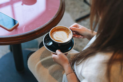 Attractive young brunette smiling woman in white casual dress with cup of coffee in hands in cafe