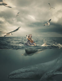 Birds flying over girl with violin sitting in sea against sky