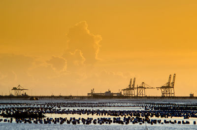 Silhouette cranes at harbor against orange sky during sunset