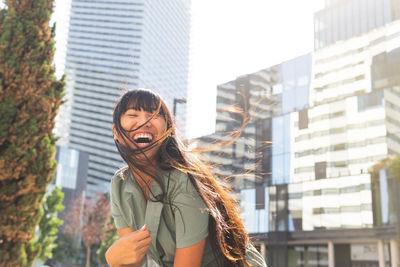 From below happy young asian female with long hair smiling with eyes closed while sitting against modern skyscrapers on sunny day on city street