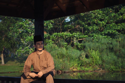 Portrait of young man sitting by plants against trees