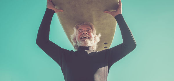 Low angle view of cheerful man carrying surfboard against clear sky