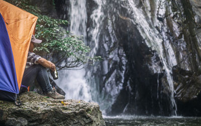 Male tourist sitting in tent and drinking coffee at the waterfall