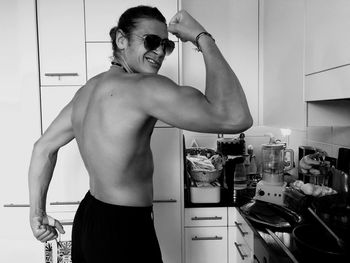 Shirtless young man flexing muscles while standing in kitchen at home