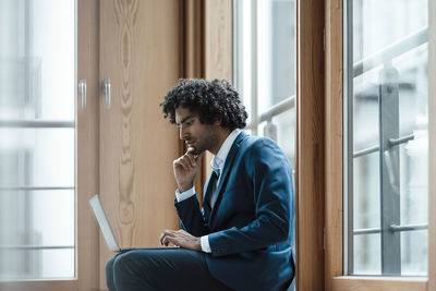 Young male entrepreneur using laptop while sitting against window at workplace