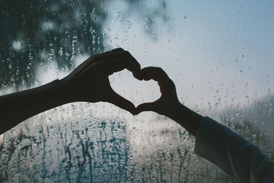 Cropped hands gesturing heart shape against wet window