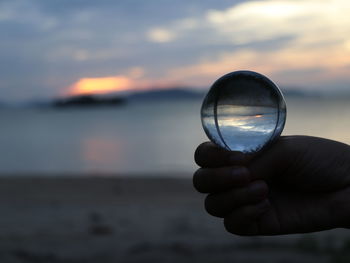 Close-up of hand holding crystal ball against sea during sunset