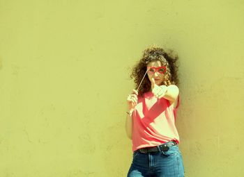 Portrait of young woman holding eye mask prop while standing against wall