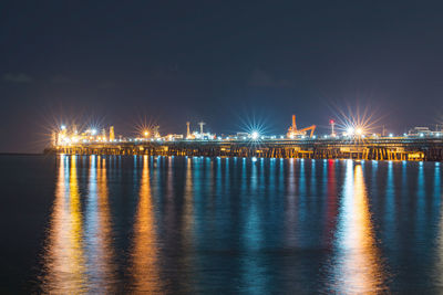 Illuminated factory by sea against sky at night