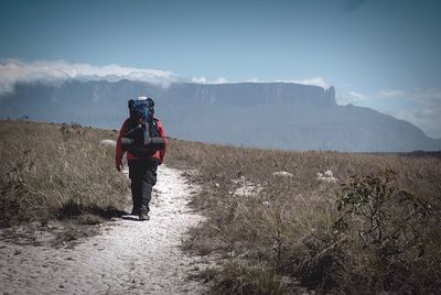 Rear view of hiker walking on dirt road amidst field towards mountains