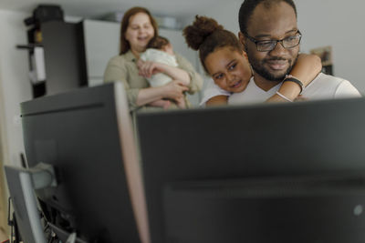 Smiling man working through computer with daughter embracing from behind