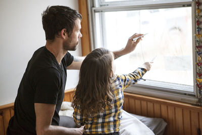 Father and son drawing on window glass while kneeling on bed at home