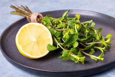 Bunch of sprigs of fresh mint and lemon lies on black ceramic plate on blue background.