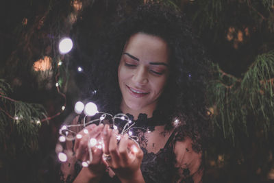 Young woman holding illuminated string lights by trees at park