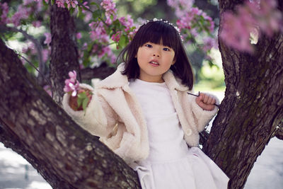 Korean girl in a white light fur coat and a headband sitting on a tree branch in a garden