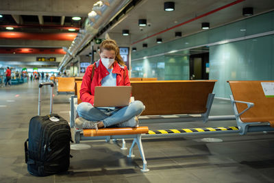 Focused female in face mask traveler typing on modern netbook while sitting near suitcase in light modern airport terminal before departure during coronavirus pandemic