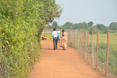 Rear view of man and woman walking on footpath amidst fence and trees