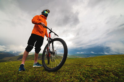 Man in a helmet and protection stands with a bicycle on a green hill among the mountains during a