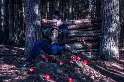 Full length of young woman holding decoration while sitting by apples in forest
