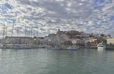 Sailboats in city on sea against cloudy sky