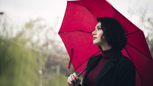 Woman looking away holding umbrella standing against sky