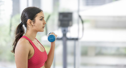 Young focused female athlete performing a bicep curl with a blue dumbbell in a bright gym.