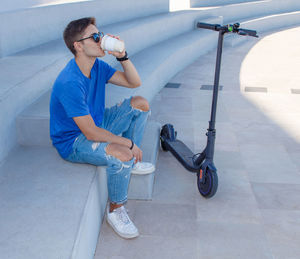 Young caucasian man in blue t-shirt sitting outdoor on stone stairs near electric scooter 