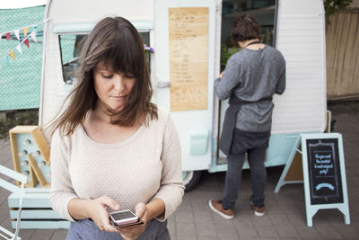 Female owner using smart phone outside food truck while colleague working in background