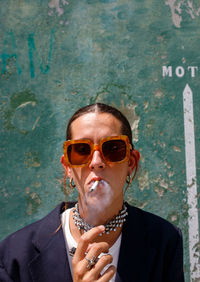 Self assured adult female with dark hair in stylish outfit and sunglasses smoking cigarette and looking at camera on street near shabby wall on sunny day