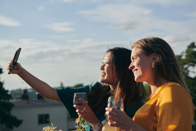 Cheerful female friends taking selfie with mobile phone on terrace during sunny day