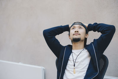 Thoughtful male computer programmer sitting with hands behind head against beige wall in office